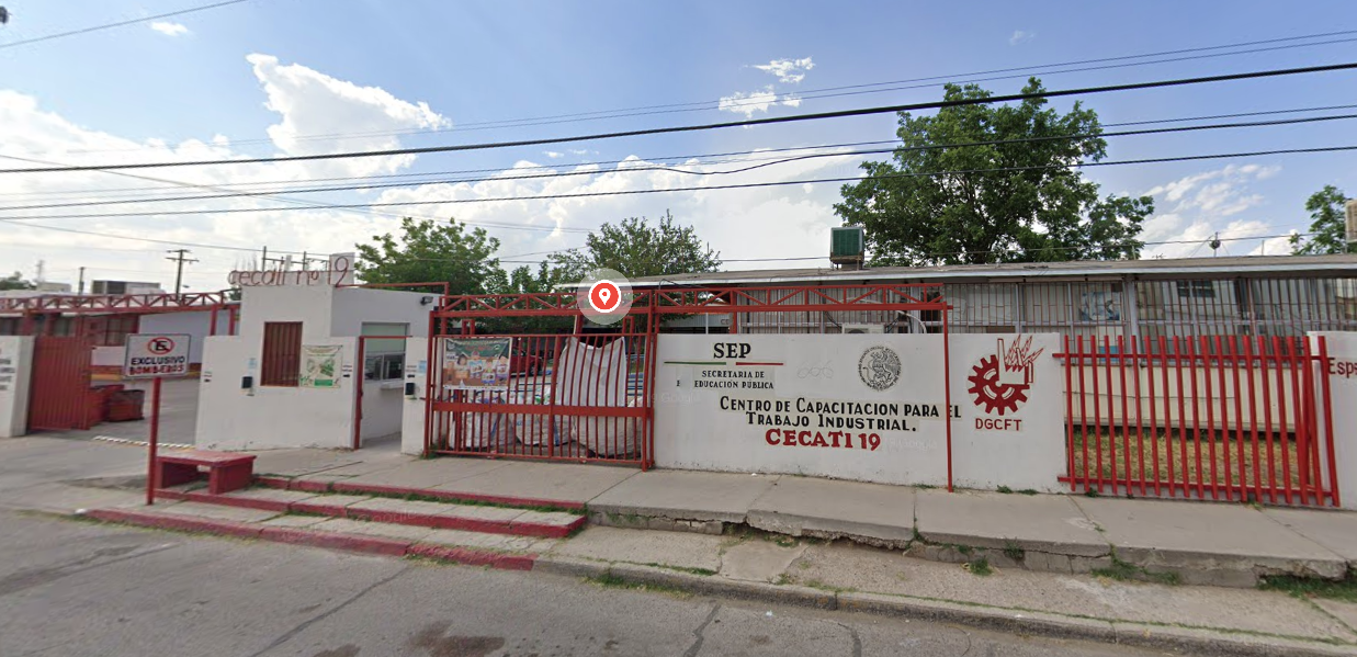 Fotografía en la que se muestra la entrada del plantel, conformada por muros blancos intercalados con rejas rojas. En uno de los muros, en letras negras y rojas dice CENTRO DE CAPACITACIÓN PARA EL TRABAJO INDUSTRIAL, CECATI 19.
